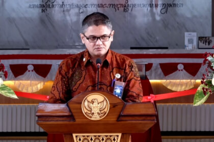 Man wearing batik shirt speaks at a wooden podium with a national emblem, flanked by flower arrangements at a formal event.