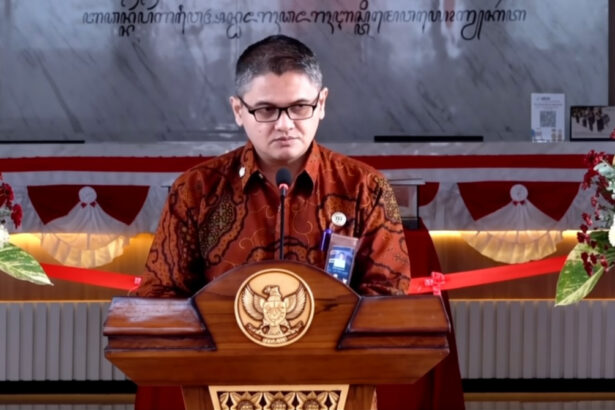 Man wearing batik shirt speaks at a wooden podium with a national emblem, flanked by flower arrangements at a formal event.