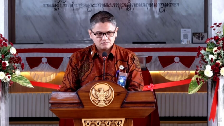 Man wearing batik shirt speaks at a wooden podium with a national emblem, flanked by flower arrangements at a formal event.