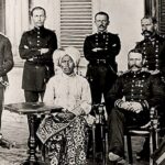 Group of uniformed military officers surrounding a central seated African woman in a turban and patterned dress; a colonial-era portrait.