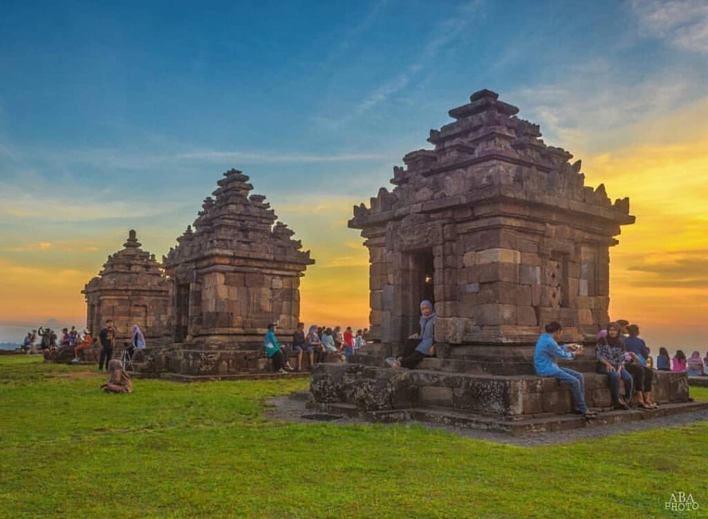 Happy Weekend😊Loc- Candi Ijo Prambananphoyo by andreasbagas_acandiijo jogja24jam - Mabur.co Tourists sit and explore a historic stone temple complex at sunset, with a grassy foreground and blue-to-orange sky.