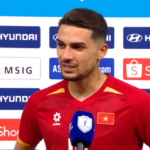 Male soccer player in a red jersey giving an interview, standing in front of sponsor boards with Hyundai and Shopee logos, blue mic in hand.