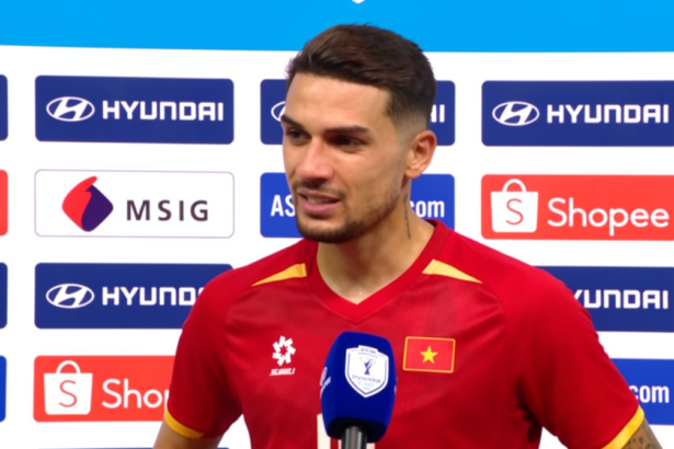 Male soccer player in a red jersey giving an interview, standing in front of sponsor boards with Hyundai and Shopee logos, blue mic in hand.
