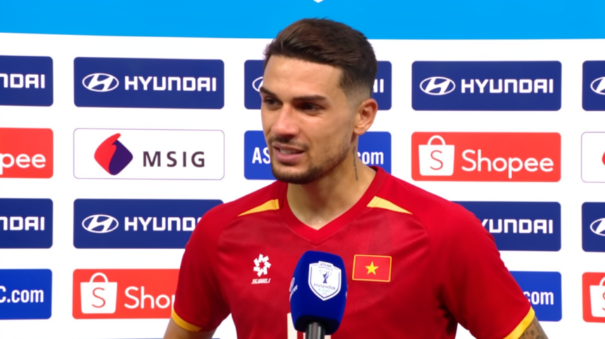 Male soccer player in a red jersey giving an interview, standing in front of sponsor boards with Hyundai and Shopee logos, blue mic in hand.