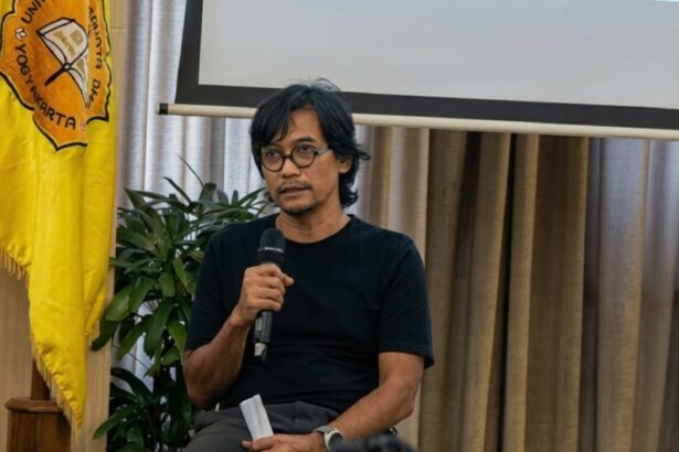 Man in a black t-shirt speaking into a handheld microphone at a panel or presentation, with a yellow flag and a projector screen behind him.