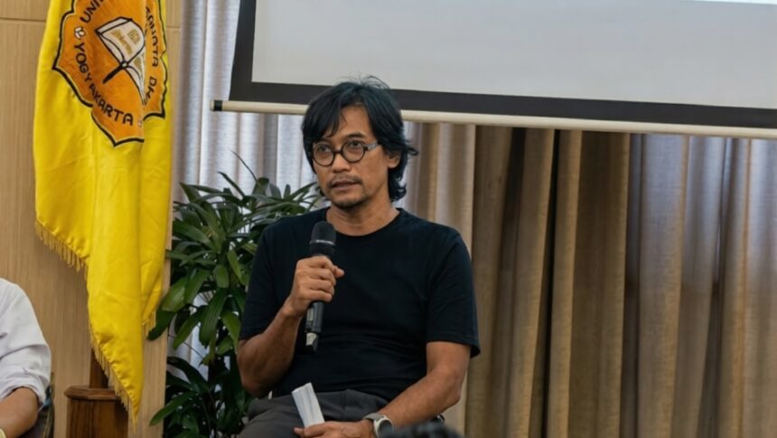 Man in a black t-shirt speaking into a handheld microphone at a panel or presentation, with a yellow flag and a projector screen behind him.