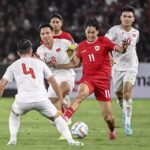 Soccer match in progress: a player in red (number 11) kicks the ball while white-jersey opponents converge.