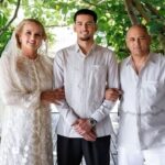Bride in lace wedding gown and veil, groom in a light shirt, standing arm-in-arm with a man in white, on a leafy veranda.