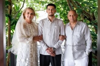Bride in lace wedding gown and veil, groom in a light shirt, standing arm-in-arm with a man in white, on a leafy veranda.