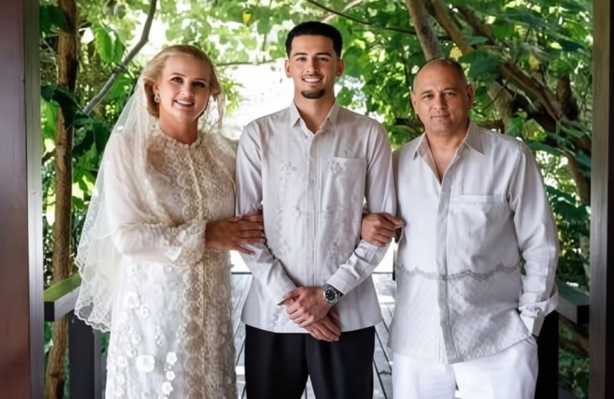Bride in lace wedding gown and veil, groom in a light shirt, standing arm-in-arm with a man in white, on a leafy veranda.