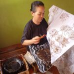 A woman works on a fabric on a loom, painting or applying wax to create batik-like circular motifs on a white cloth.
