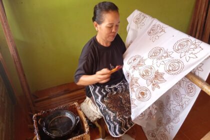 A woman works on a fabric on a loom, painting or applying wax to create batik-like circular motifs on a white cloth.
