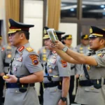 Two uniformed police officers in a ceremonial setting; one officer presents a booklet to another while several colleagues stand in the background.