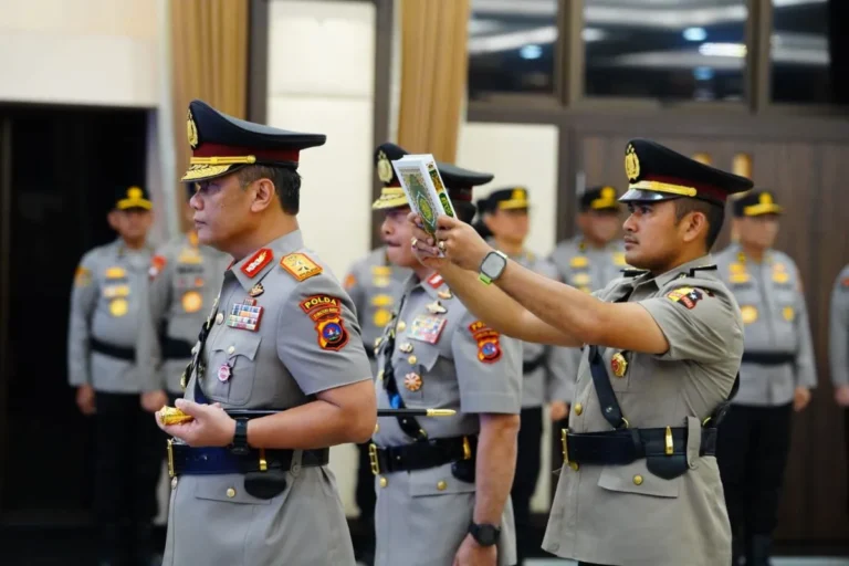 Two uniformed police officers in a ceremonial setting; one officer presents a booklet to another while several colleagues stand in the background.