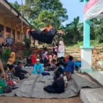 Group of children sitting on mats outside a school building, supervised by adults on a sunny day in a courtyard area.