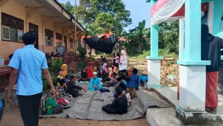 Group of children sitting on mats outside a school building, supervised by adults on a sunny day in a courtyard area.