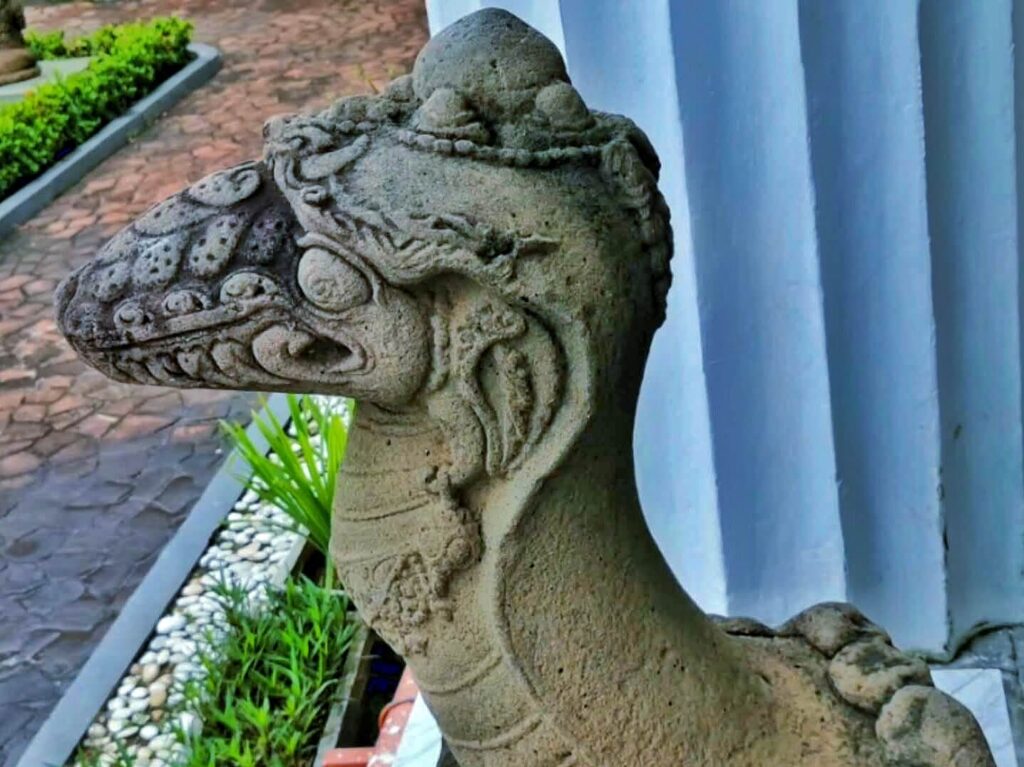 Stone dragon statue head in profile, weathered gray, with carved scales and ornamental details outdoors by a blue wall and garden path