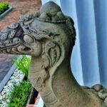 Stone dragon statue head in profile, weathered gray, with carved scales and ornamental details outdoors by a blue wall and garden path