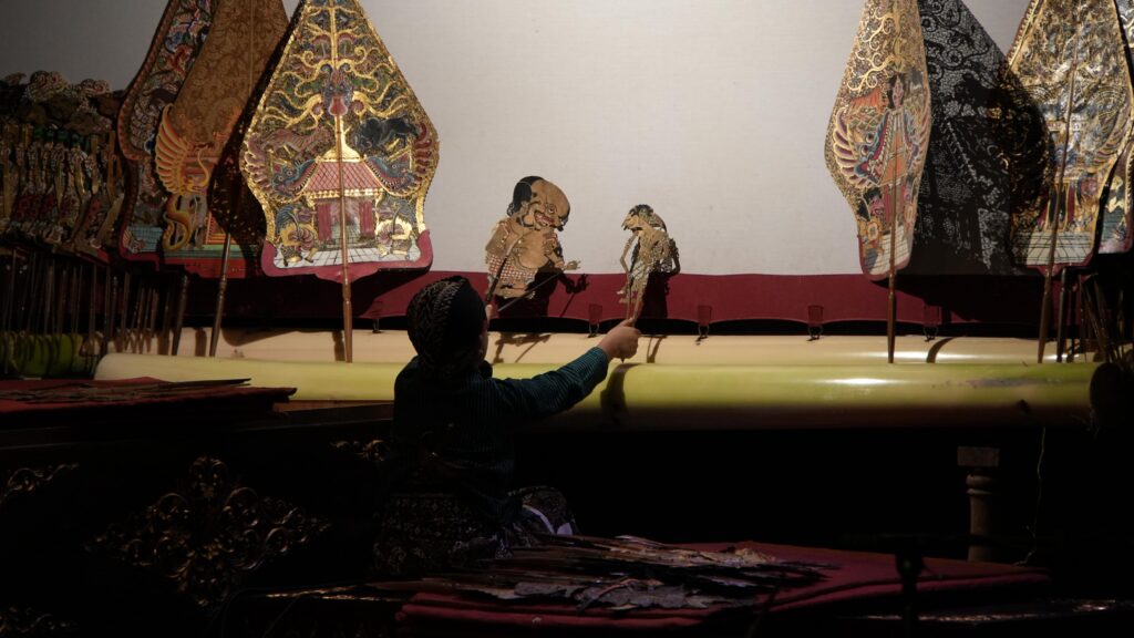 A puppeteer in a dark outfit operates two ornate traditional puppets on a dimly lit stage with decorative backdrops.