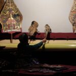 A puppeteer in a dark outfit operates two ornate traditional puppets on a dimly lit stage with decorative backdrops.