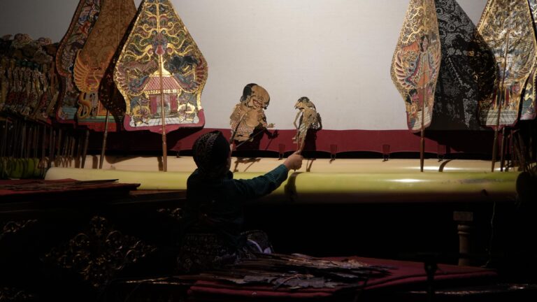 A puppeteer in a dark outfit operates two ornate traditional puppets on a dimly lit stage with decorative backdrops.