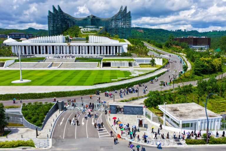 Aerial view of a modern campus-like complex with a curved road, tiered terraces, and a bright green lawn filled with people walking.