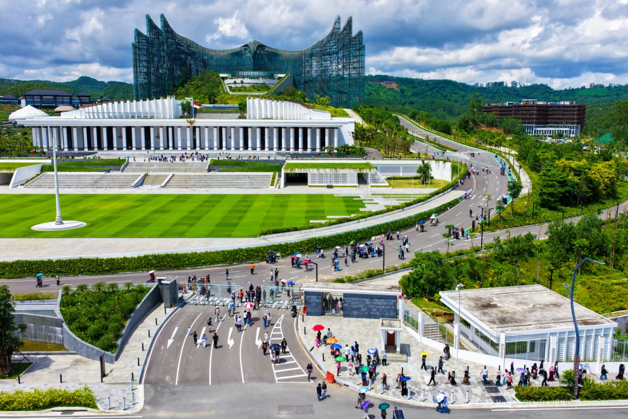 Aerial view of a modern campus-like complex with a curved road, tiered terraces, and a bright green lawn filled with people walking.