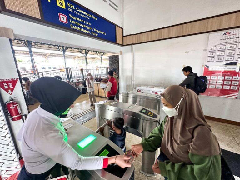 People pass through metal turnstiles at a station gate, with a blue sign overhead and passengers waiting nearby.