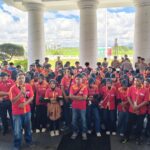 Large group of students in red and yellow uniforms gathers under a columned pavilion for an event outside, with a grassy field in the background.