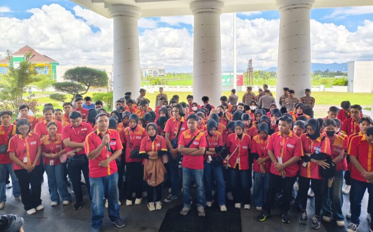 Large group of students in red and yellow uniforms gathers under a columned pavilion for an event outside, with a grassy field in the background.