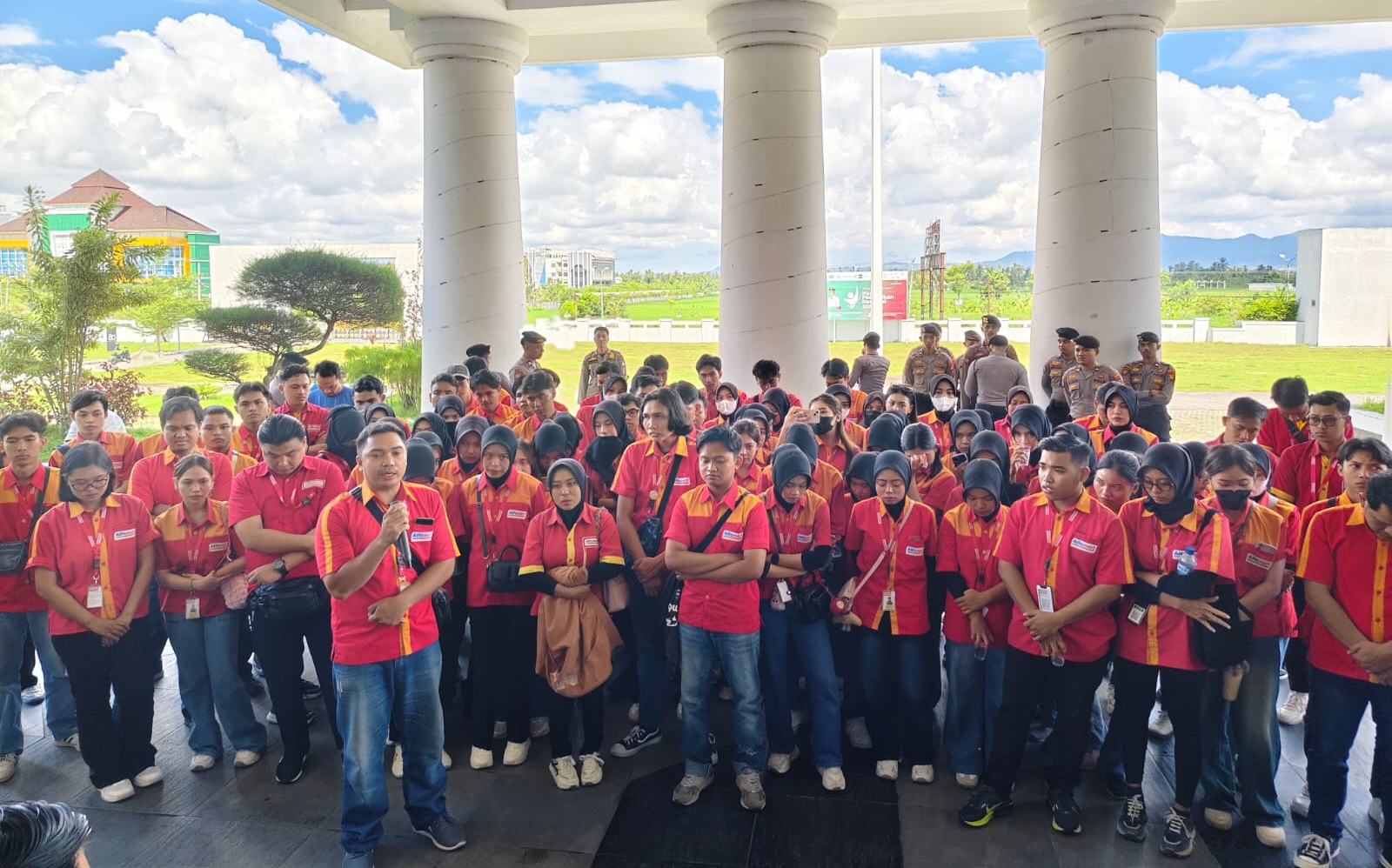Large group of students in red and yellow uniforms gathers under a columned pavilion for an event outside, with a grassy field in the background.