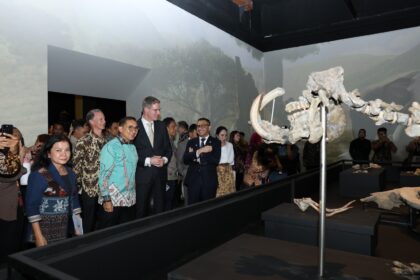 Group of diverse visitors in formal and traditional attire standing along a railing, looking at a large fossil skeleton exhibit in a museum.