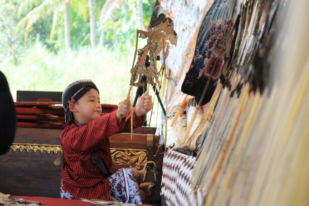 Young child in a red striped shirt and headscarf holds intricate wooden puppets at a stall with colorful fans on display behind him.