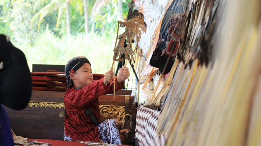 Young child in a red striped shirt and headscarf holds intricate wooden puppets at a stall with colorful fans on display behind him.