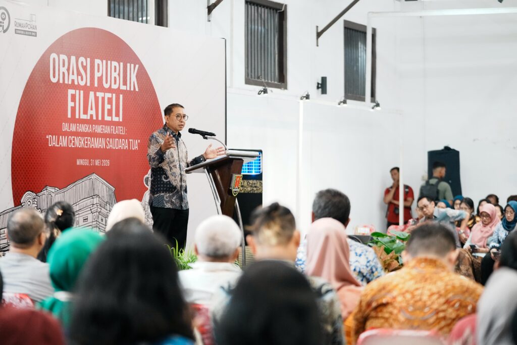 Man at a lectern delivering a talk to a seated audience, with a large red banner reading'ORASI PUBLIK FILATELI' behind him.