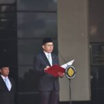 Indonesian official in a suit and peci delivers a speech at a podium with a national emblem, microphone in front, a security guard nearby.
