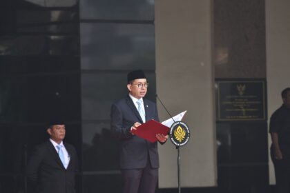 Indonesian official in a suit and peci delivers a speech at a podium with a national emblem, microphone in front, a security guard nearby.