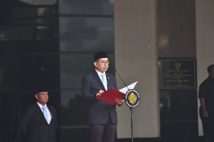 Indonesian official in a suit and peci delivers a speech at a podium with a national emblem, microphone in front, a security guard nearby.