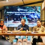 Three speakers sit at a wooden table with snacks, microphones, and bottles; a large screen behind them shows an Indonesian event banner in a cafe setting.