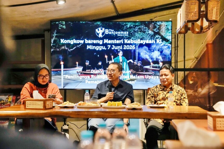 Three speakers sit at a wooden table with snacks, microphones, and bottles; a large screen behind them shows an Indonesian event banner in a cafe setting.