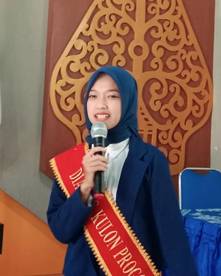 Young woman in a blue hijab and blazer speaks into a handheld microphone at a formal event, wearing a red sash with gold trim.