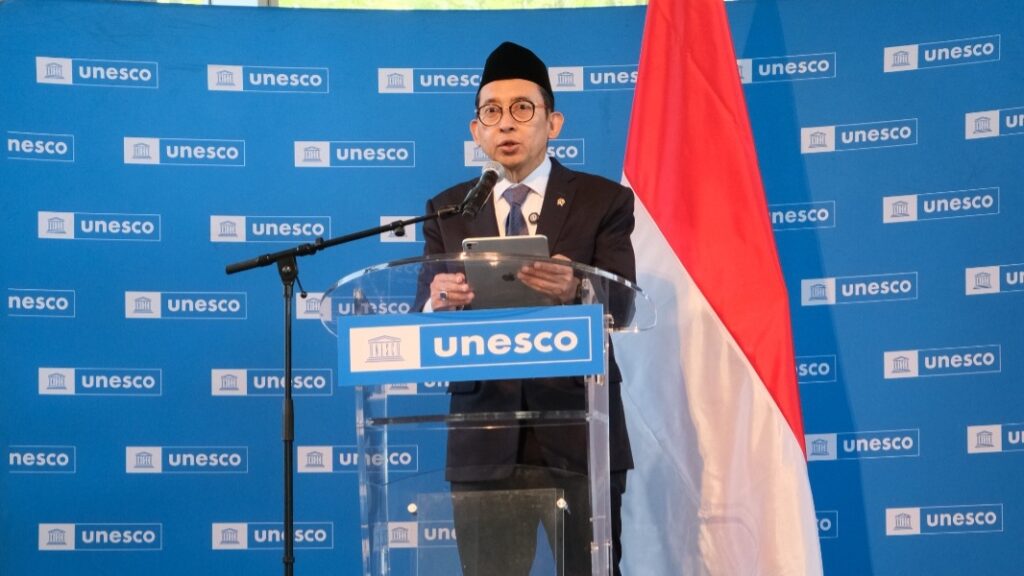 IMG-20260609-WA0001 - Mabur.co Man in a suit and glasses wearing a traditional cap speaks at a UNESCO podium, holding a tablet, with a blue UNESCO backdrop and the Indonesian flag beside him.