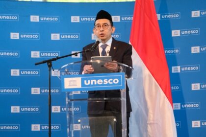Man in a suit and glasses wearing a traditional cap speaks at a UNESCO podium, holding a tablet, with a blue UNESCO backdrop and the Indonesian flag beside him.