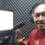 Smiling man wearing headphones in a soundproof recording booth, waving toward a microphone and computer monitor beside him.