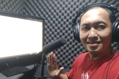 Smiling man wearing headphones in a soundproof recording booth, waving toward a microphone and computer monitor beside him.