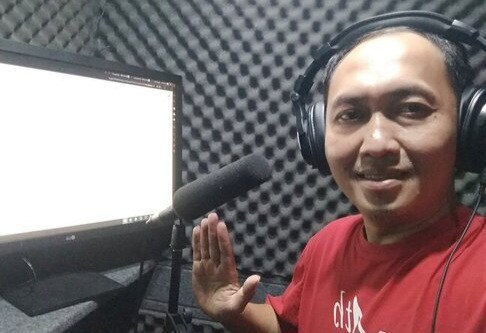 IMG-20260611-WA0001 - Mabur.co Smiling man wearing headphones in a soundproof recording booth, waving toward a microphone and computer monitor beside him.