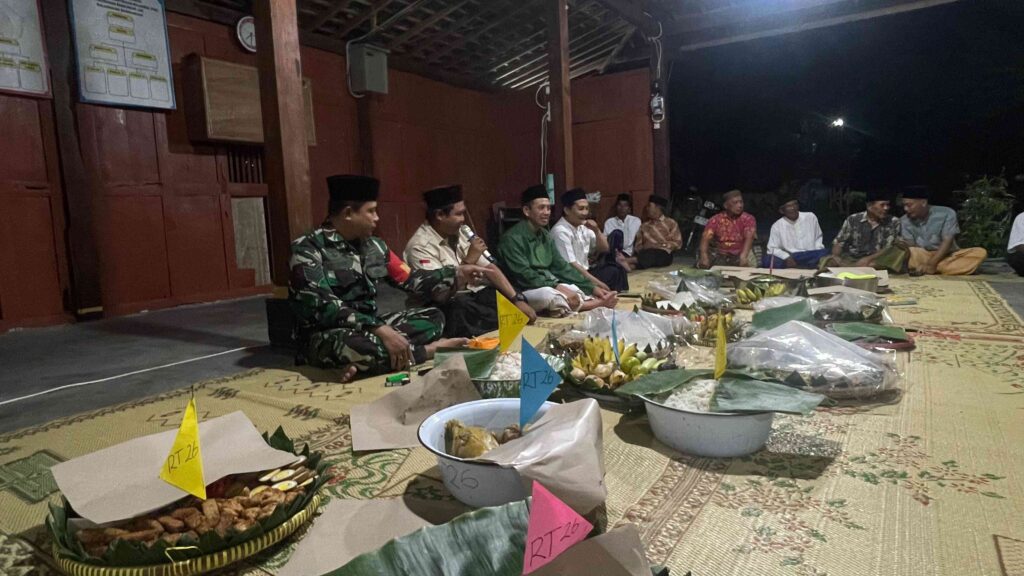 IMG-20260616-WA0002 - Mabur.co Group of men in traditional clothing and a soldier sit on a mat at night, surrounding baskets of ceremonial food offerings.