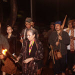 Group of people in traditional clothing walking at night, one man lighting a torch while others carry staffs and instruments.