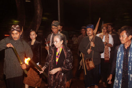 Group of people in traditional clothing walking at night, one man lighting a torch while others carry staffs and instruments.