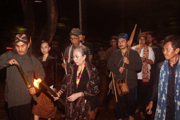Group of people in traditional clothing walking at night, one man lighting a torch while others carry staffs and instruments.